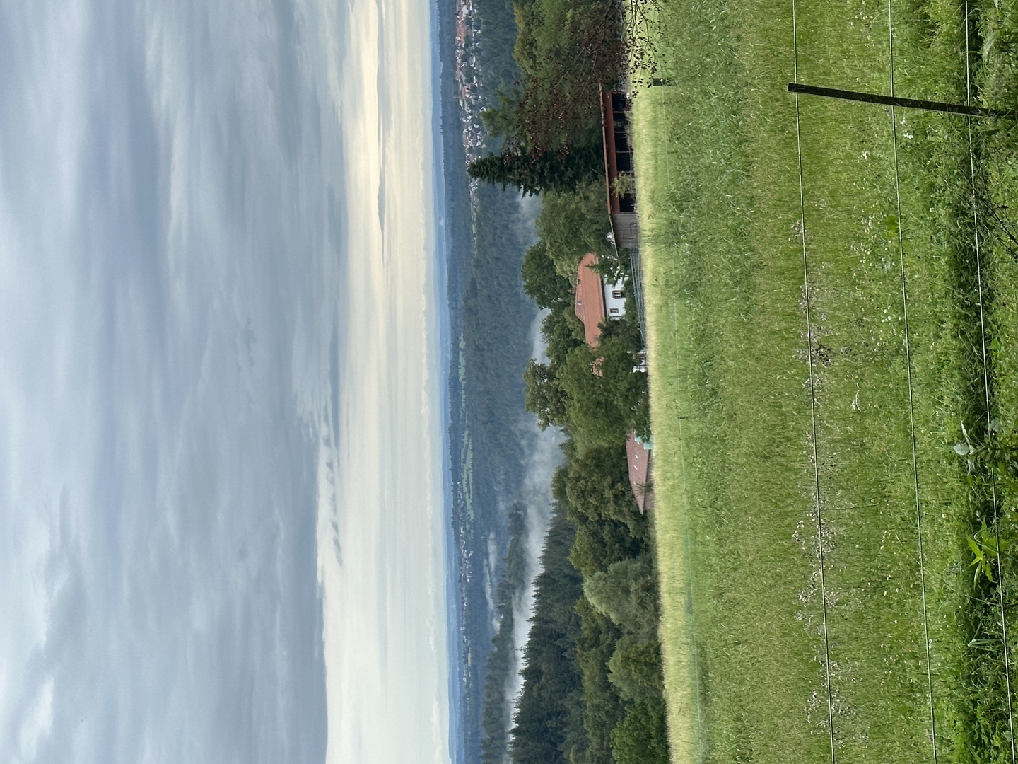 Landschaft mit grünen Wiesen, Bäumen, Häusern und einem ruhigen Gewässer im Hintergrund.