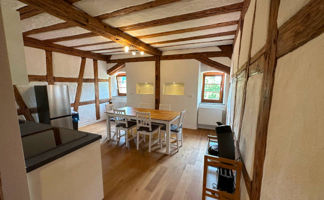 Ein helles Wohnzimmer mit Fachwerkwänden, Holzbalken an der Decke, einem Esstisch mit Stühlen, einer Küchenzeile und großen Fenstern.