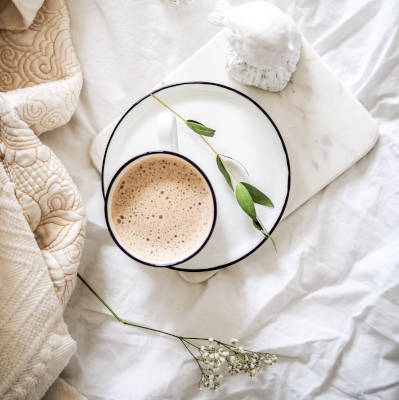 Eine Tasse mit cremigem Getränk auf einem weißen Teller, dekoriert mit einer grünen Blattspitze, steht auf einem Bett mit cremigen Kissen und einer weißen Bettdecke.